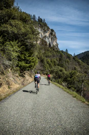 Cyclists riding along rugged coastal cliffs with the Mediterranean Sea sparkling beneath, under a clear blue sky.