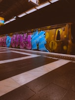 A subway platform with a train covered in colorful graffiti art. The platform floor is tiled with a combination of dark and light tiles, and there are station signs with numbers overhead. The graffiti on the train includes shades of blue, pink, and yellow, creating a vibrant urban feel.