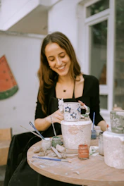 a woman sitting at a table painting a vase