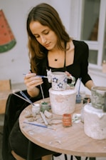 A woman is focused on painting a ceramic mug with a floral design. She sits at a round table cluttered with paint jars, brushes, and clay tools. The setting appears to be a studio or workshop with a calm, creative atmosphere.