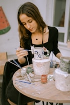 A woman is focused on painting a ceramic mug with a floral design. She sits at a round table cluttered with paint jars, brushes, and clay tools. The setting appears to be a studio or workshop with a calm, creative atmosphere.