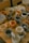 A collection of handmade ceramic mugs and bowls displayed on a wooden table. The items appear to be uniquely designed with various patterns and colors, including stripes, hearts, and floral motifs. The ceramics are in shades of orange, gray, and beige, each with a rustic and artistic handmade appearance.