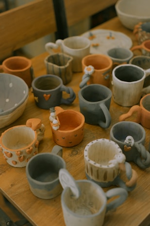A collection of handmade ceramic mugs and bowls displayed on a wooden table. The items appear to be uniquely designed with various patterns and colors, including stripes, hearts, and floral motifs. The ceramics are in shades of orange, gray, and beige, each with a rustic and artistic handmade appearance.