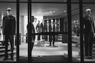 A black and white image of a high-end clothing store display featuring mannequins dressed in formal attire. The mannequins are positioned both in the storefront window and inside the shop, showcasing suits and elegant fashion items. A person is visible near the entrance, partially obscured by the window frame, adding an element of mystery.