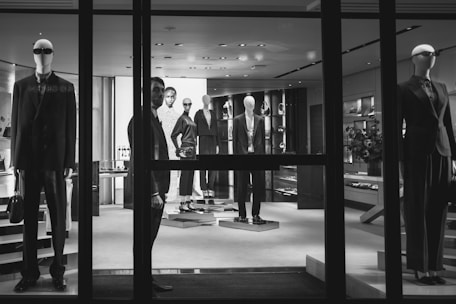 A black and white image of a high-end clothing store display featuring mannequins dressed in formal attire. The mannequins are positioned both in the storefront window and inside the shop, showcasing suits and elegant fashion items. A person is visible near the entrance, partially obscured by the window frame, adding an element of mystery.