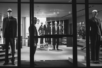 A black and white image of a high-end clothing store display featuring mannequins dressed in formal attire. The mannequins are positioned both in the storefront window and inside the shop, showcasing suits and elegant fashion items. A person is visible near the entrance, partially obscured by the window frame, adding an element of mystery.