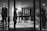 A black and white image of a high-end clothing store display featuring mannequins dressed in formal attire. The mannequins are positioned both in the storefront window and inside the shop, showcasing suits and elegant fashion items. A person is visible near the entrance, partially obscured by the window frame, adding an element of mystery.