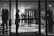 A black and white image of a high-end clothing store display featuring mannequins dressed in formal attire. The mannequins are positioned both in the storefront window and inside the shop, showcasing suits and elegant fashion items. A person is visible near the entrance, partially obscured by the window frame, adding an element of mystery.