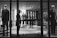 A black and white image of a high-end clothing store display featuring mannequins dressed in formal attire. The mannequins are positioned both in the storefront window and inside the shop, showcasing suits and elegant fashion items. A person is visible near the entrance, partially obscured by the window frame, adding an element of mystery.