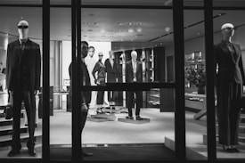 A black and white image of a high-end clothing store display featuring mannequins dressed in formal attire. The mannequins are positioned both in the storefront window and inside the shop, showcasing suits and elegant fashion items. A person is visible near the entrance, partially obscured by the window frame, adding an element of mystery.