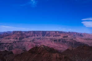 A vast, expansive view of the Grand Canyon under a clear blue sky. The canyon features layered red and brown rock formations displaying varying shades and textures.