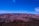 A vast, expansive view of the Grand Canyon under a clear blue sky. The canyon features layered red and brown rock formations displaying varying shades and textures.
