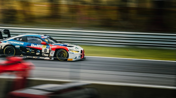 A race car is speeding along a track, creating a motion blur effect. The car has a multi-colored body with prominent sponsorship decals and the number 8 on its side. The background shows a metal guardrail and blurred natural scenery, emphasizing the high speed.