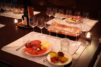 A table set with wine glasses and a variety of appetizers on white placemats. The appetizers include cured meats, cheese, and vegetables on plates. There are lit candles on the table and a bottle of wine in the center.