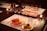 A table set with wine glasses and a variety of appetizers on white placemats. The appetizers include cured meats, cheese, and vegetables on plates. There are lit candles on the table and a bottle of wine in the center.