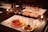 A table set with wine glasses and a variety of appetizers on white placemats. The appetizers include cured meats, cheese, and vegetables on plates. There are lit candles on the table and a bottle of wine in the center.