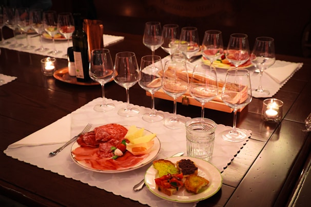 A table set with wine glasses and a variety of appetizers on white placemats. The appetizers include cured meats, cheese, and vegetables on plates. There are lit candles on the table and a bottle of wine in the center.