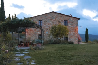 A rustic stone house surrounded by greenery, with a canopy-covered seating area featuring wooden benches and a table. The setting is serene, with a manicured lawn and a few small trees. The sky is overcast, casting a soft, diffused light over the landscape.