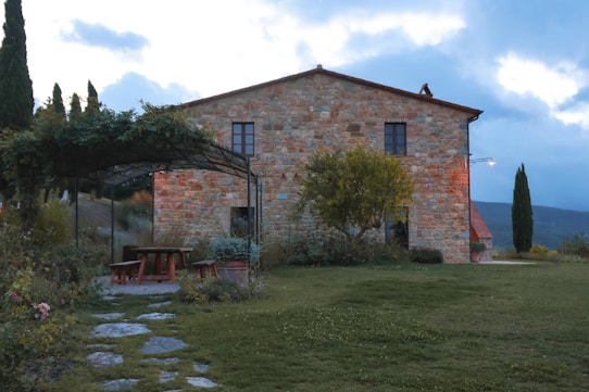 A rustic stone house surrounded by greenery, with a canopy-covered seating area featuring wooden benches and a table. The setting is serene, with a manicured lawn and a few small trees. The sky is overcast, casting a soft, diffused light over the landscape.