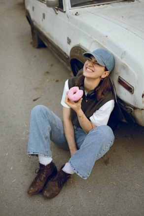 A person wearing a blue cap and jeans is sitting on the ground next to an old, white car. They are smiling broadly while holding a pink frosted donut in one hand. The person is dressed casually, with brown boots and a dark vest over a white shirt.