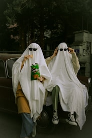 Two people are dressed in humorous ghost costumes made from white sheets and sunglasses while sitting on an old car. One is holding a bag of Doritos and eating a chip. The setting is outdoors, surrounded by trees and a building.