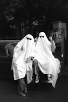 Three people are dressed up as ghosts, wearing white sheets and black sunglasses, sitting on the hood of a vintage car. The background is wooded, with some elements of a building visible.