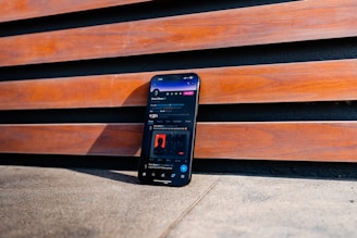 A smartphone is positioned upright against a wooden slat wall. The phone screen displays a social media app interface. The wooden slats have a warm, reddish-brown color, and the ground appears to be a rough concrete surface.