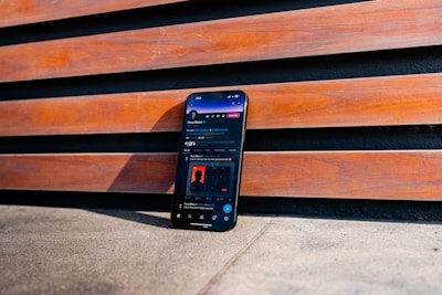 A smartphone is positioned upright against a wooden slat wall. The phone screen displays a social media app interface. The wooden slats have a warm, reddish-brown color, and the ground appears to be a rough concrete surface.