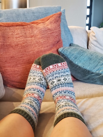 A pair of feet wearing patterned socks is resting on a couch. The socks feature a knit design with geometric patterns in colors such as red, blue, white, and green. Behind the feet, there are several cushions in earthy tones, including a muted orange, light blue, and dark teal.