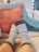 A pair of feet wearing patterned socks is resting on a couch. The socks feature a knit design with geometric patterns in colors such as red, blue, white, and green. Behind the feet, there are several cushions in earthy tones, including a muted orange, light blue, and dark teal.