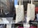 Two fabric tote bags are on display in a shop window. The bag on the left features a pattern of hand-drawn middle fingers, while the bag on the right displays a pattern of hand-drawn breasts. The background shows a street with cars and buildings, and the reflection of the cityscape can be seen on the glass.