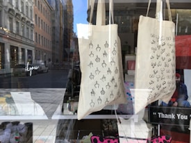 Two fabric tote bags are on display in a shop window. The bag on the left features a pattern of hand-drawn middle fingers, while the bag on the right displays a pattern of hand-drawn breasts. The background shows a street with cars and buildings, and the reflection of the cityscape can be seen on the glass.