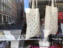 Two fabric tote bags are on display in a shop window. The bag on the left features a pattern of hand-drawn middle fingers, while the bag on the right displays a pattern of hand-drawn breasts. The background shows a street with cars and buildings, and the reflection of the cityscape can be seen on the glass.