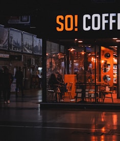A cozy coffee shop with warm lighting is visible through a large front window. Inside, several people are seated at tables, creating a relaxed atmosphere. Outside, it appears to be evening as the surrounding area is dimly lit, with a few people walking by.