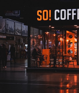 A cozy coffee shop with warm lighting is visible through a large front window. Inside, several people are seated at tables, creating a relaxed atmosphere. Outside, it appears to be evening as the surrounding area is dimly lit, with a few people walking by.