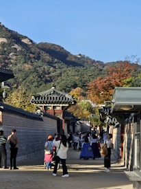A sunny day in a traditional Korean setting with tiled roofs and a stone wall. People are walking and interacting, some dressed in traditional Korean hanbok attire. The background features a forested mountain landscape with autumn leaves.