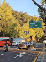 A busy urban street corner with several vehicles, including motorcycles and cars, moving on the road. There are lush trees with vibrant yellow leaves, suggesting an autumn setting. The sky is clear and blue, and traffic signs in Korean are visible, indicating directions to various locations.