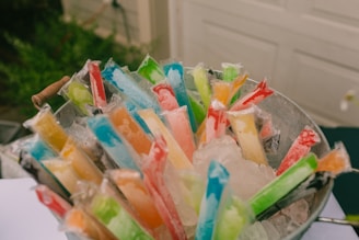 A metal bucket is filled with colorful ice pops, each wrapped in clear plastic bags. The ice pops are nestled on a bed of crushed ice, showcasing a variety of vibrant colors, including red, blue, green, orange, and yellow.