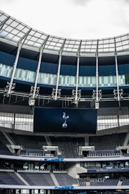 A modern stadium interior featuring a large digital screen with a logo prominently displayed. The seating areas are empty, with dark-colored seats and some structural elements like beams and glass walls visible. A combination of dark blue and neutral tones are prominent.