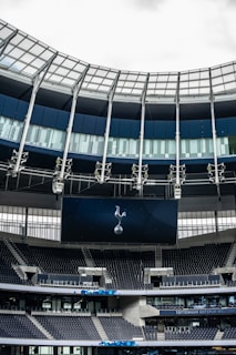 A modern stadium interior featuring a large digital screen with a logo prominently displayed. The seating areas are empty, with dark-colored seats and some structural elements like beams and glass walls visible. A combination of dark blue and neutral tones are prominent.