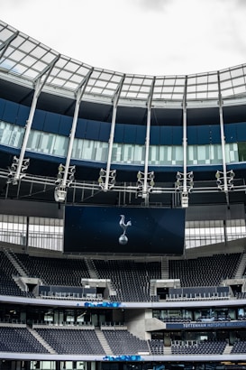 A modern stadium interior featuring a large digital screen with a logo prominently displayed. The seating areas are empty, with dark-colored seats and some structural elements like beams and glass walls visible. A combination of dark blue and neutral tones are prominent.