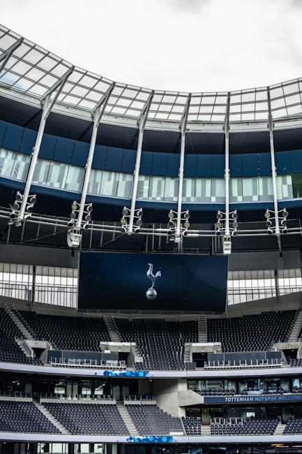 A modern stadium interior featuring a large digital screen with a logo prominently displayed. The seating areas are empty, with dark-colored seats and some structural elements like beams and glass walls visible. A combination of dark blue and neutral tones are prominent.