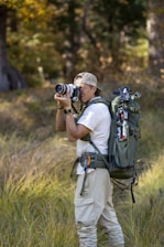 A person in outdoor attire is standing in a grassy area with trees in the background. They are holding a camera, appearing to take a photograph. A large, green backpack is strapped to their back, equipped with various outdoor gear. The scene suggests a focus on nature photography.