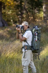 A person in outdoor attire is standing in a grassy area with trees in the background. They are holding a camera, appearing to take a photograph. A large, green backpack is strapped to their back, equipped with various outdoor gear. The scene suggests a focus on nature photography.