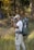 A person in outdoor attire is standing in a grassy area with trees in the background. They are holding a camera, appearing to take a photograph. A large, green backpack is strapped to their back, equipped with various outdoor gear. The scene suggests a focus on nature photography.