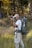 A person in outdoor attire is standing in a grassy area with trees in the background. They are holding a camera, appearing to take a photograph. A large, green backpack is strapped to their back, equipped with various outdoor gear. The scene suggests a focus on nature photography.