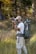 A person in outdoor attire is standing in a grassy area with trees in the background. They are holding a camera, appearing to take a photograph. A large, green backpack is strapped to their back, equipped with various outdoor gear. The scene suggests a focus on nature photography.