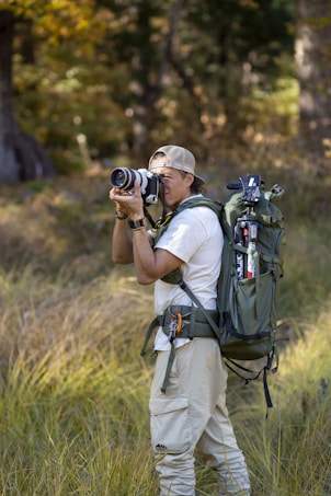 A person in outdoor attire is standing in a grassy area with trees in the background. They are holding a camera, appearing to take a photograph. A large, green backpack is strapped to their back, equipped with various outdoor gear. The scene suggests a focus on nature photography.