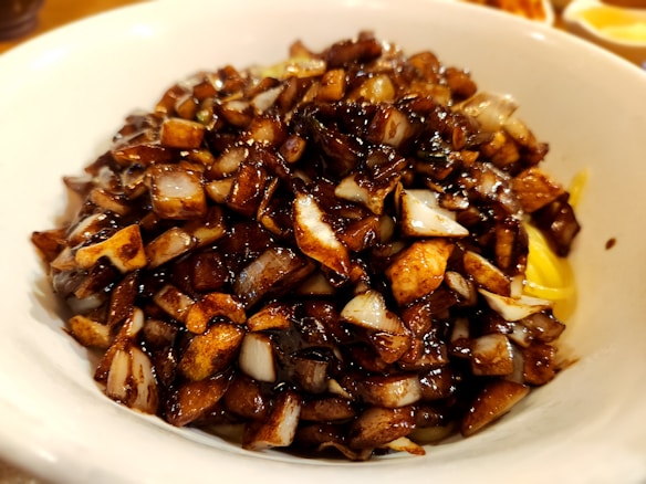 A close-up view of a bowl filled with diced vegetables, primarily onions, that appear to be cooked or sautéed and are coated with a glistening dark sauce. The bowl is white, and the background shows hints of other plateware or food items out of focus.