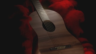 An acoustic guitar with a wooden finish is surrounded by dark shadows interspersed with vivid red smoke or fabric in the background. The lighting creates a dramatic contrast, highlighting the guitar's strings and body.